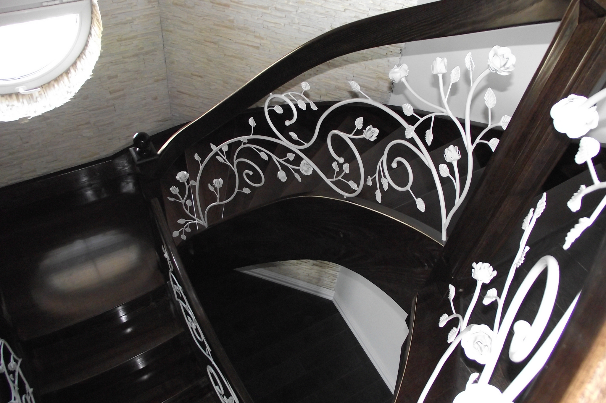 Eleganckie, ciemne schody z dekoracyjną, białą, metalową balustradą z motywem kwiatowym, oświetlone przez okrągłe okno w górze.