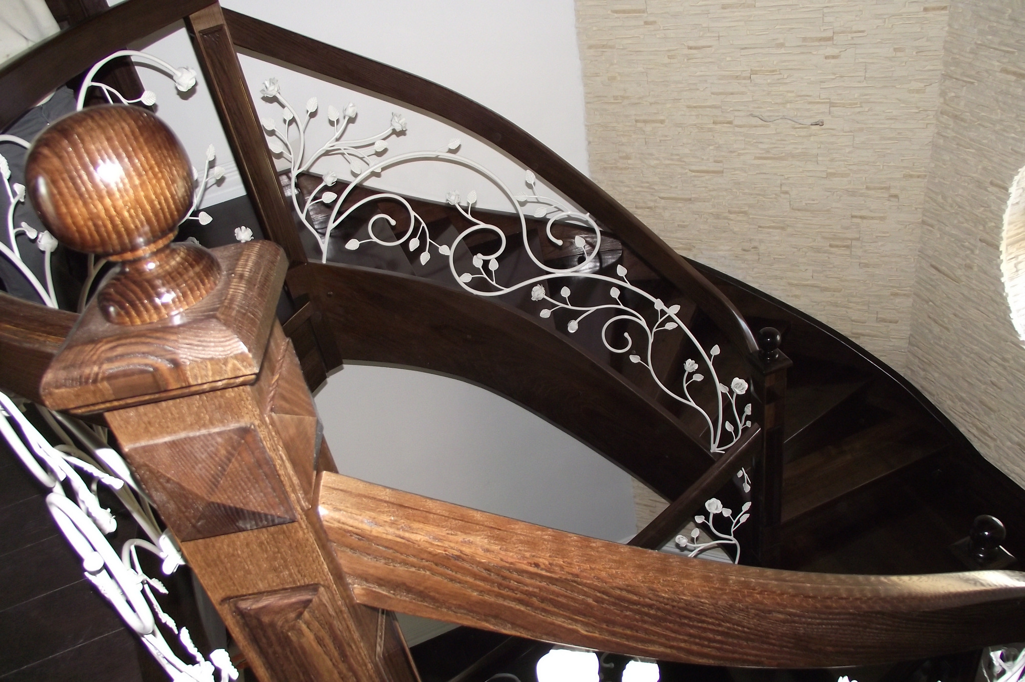 Kręte schody z ciemnego drewna z dekoracyjną, białą, metalową balustradą z motywem roślinnym, zakończone drewnianą kulą na słupku.