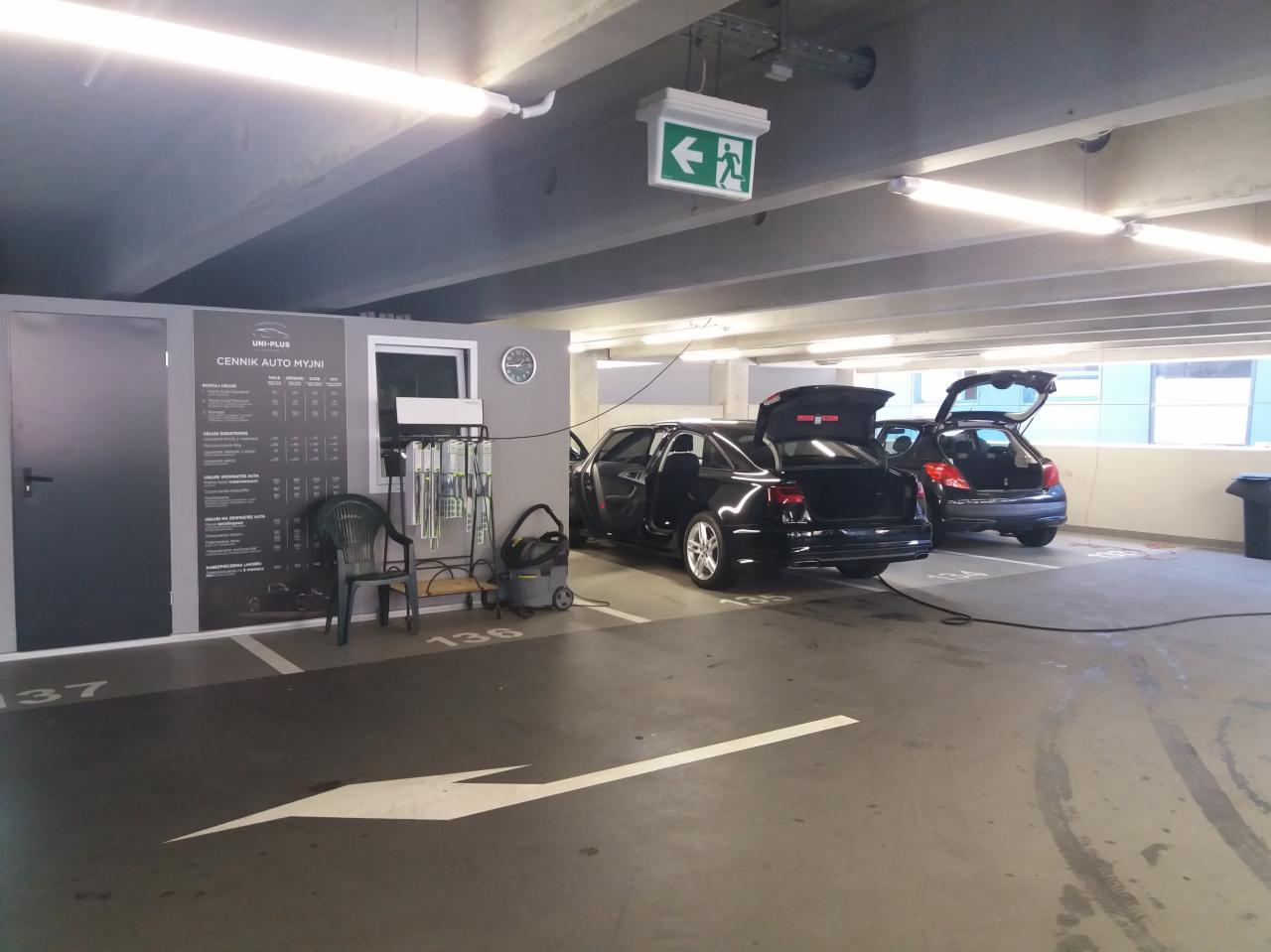 Dwa samochody z otwartymi bagażnikami na stanowisku myjni samoobsługowej w garażu podziemnym, widoczny cennik usług i sprzęt do mycia.