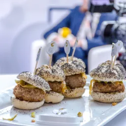 Cztery mini burgery z kotletem, ogórkiem konserwowym i bułką posypaną makiem, podane na białym talerzu z logotypem firmy na wykałaczkach, w tle rozmazana postać mężczyzny.