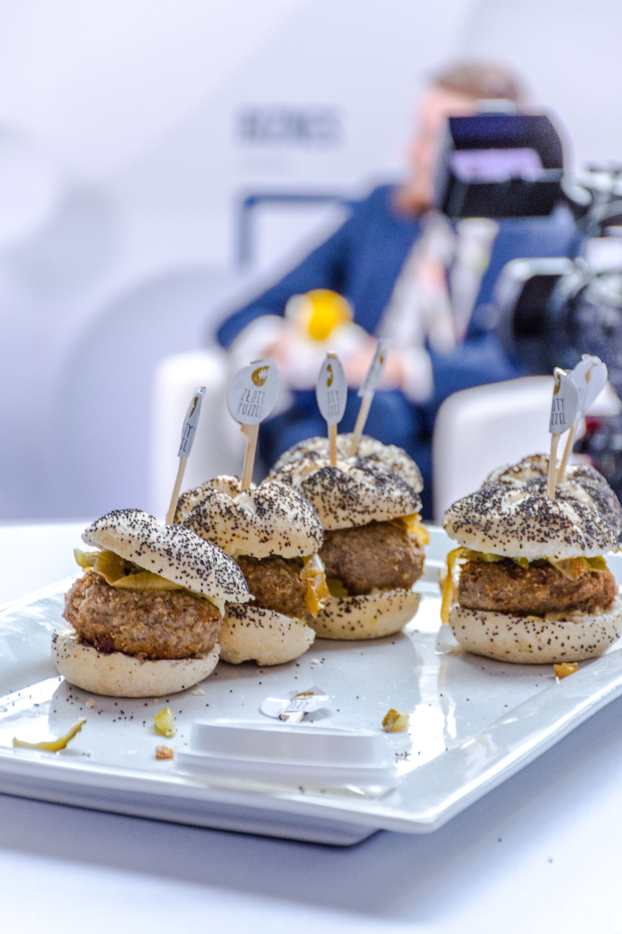 Cztery mini burgery z kotletem, ogórkiem konserwowym i bułką posypaną makiem, podane na białym talerzu z logotypem firmy na wykałaczkach, w tle rozmazana postać mężczyzny.
