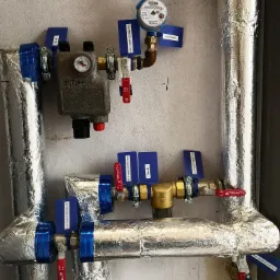 Węzeł cieplny z izolowanymi rurami, manometrem i zaworami oznaczonymi etykietami, zamontowany na szarej ścianie.