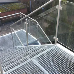 Zewnętrzne metalowe schody kratowe z balustradą ze stali nierdzewnej i szkła, widok z góry, w tle zieleń i budynek gospodarczy.