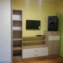 Zabudowa ściany z szafą przesuwną, regałem i szafką RTV w kolorach beżu i bieli, wykonana na zamówienie w Kole, z widocznym głośnikiem i telewizorem na ścianie.