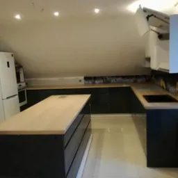 Nowoczesna kuchnia w szaro-białej tonacji na poddaszu ze skośnym sufitem, z wyspą kuchenną i blatem z jasnego drewna oraz czarnymi frontami szafek.