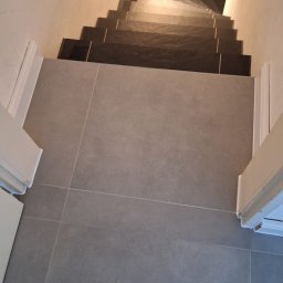 Usługi ogólnobudowlane - Szare płytki podłogowe i ciemne płytki schodowe, widok z góry. Widoczne fragmenty drzwi po bokach i but w rogu. Minimalistyczny design.