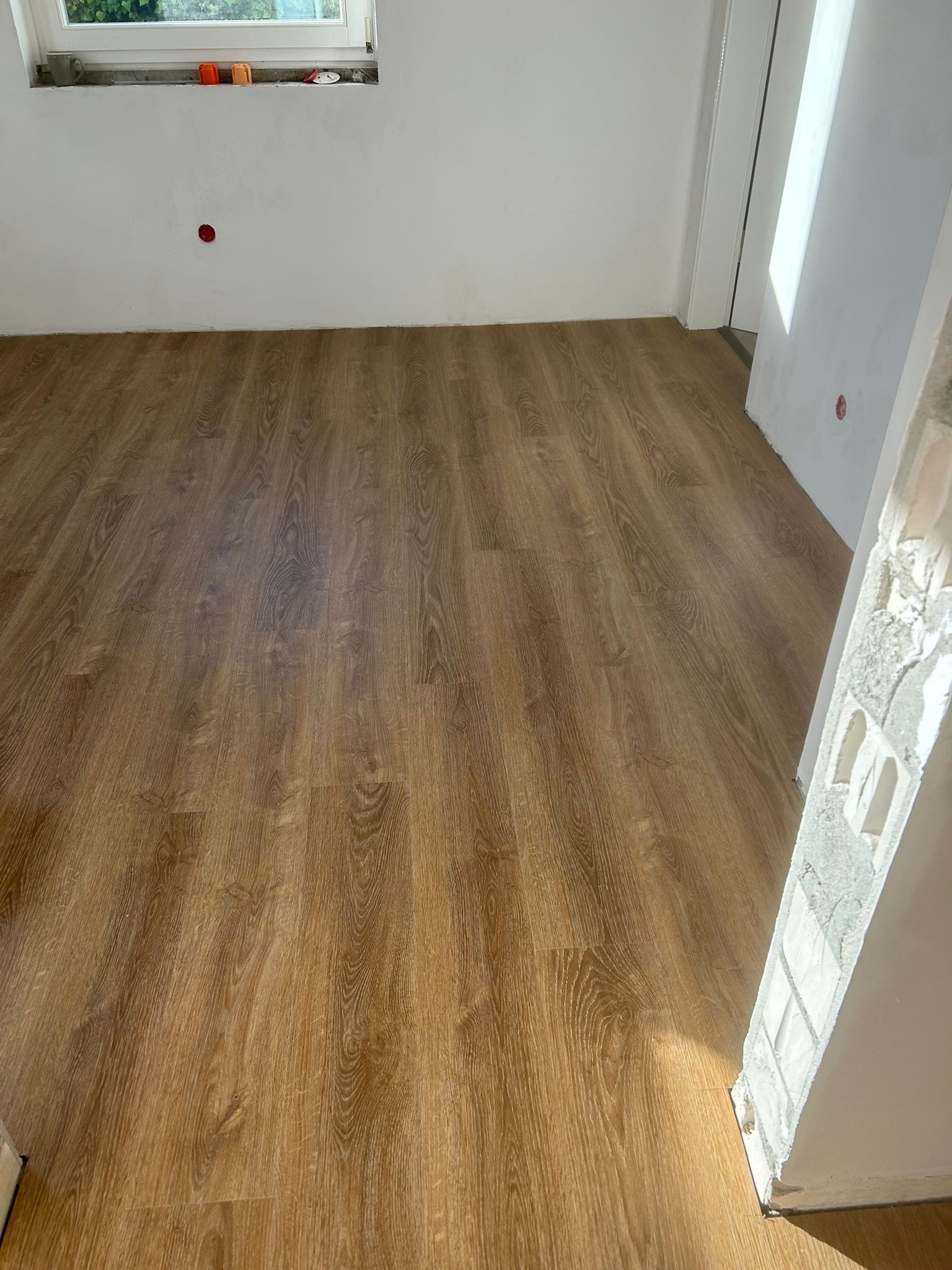 Nowo ułożone panele podłogowe o wzorze drewna w jasnym pomieszczeniu z oknem. Fragment ściany z niedokończonym wykończeniem. Widok z góry, naturalne światło.