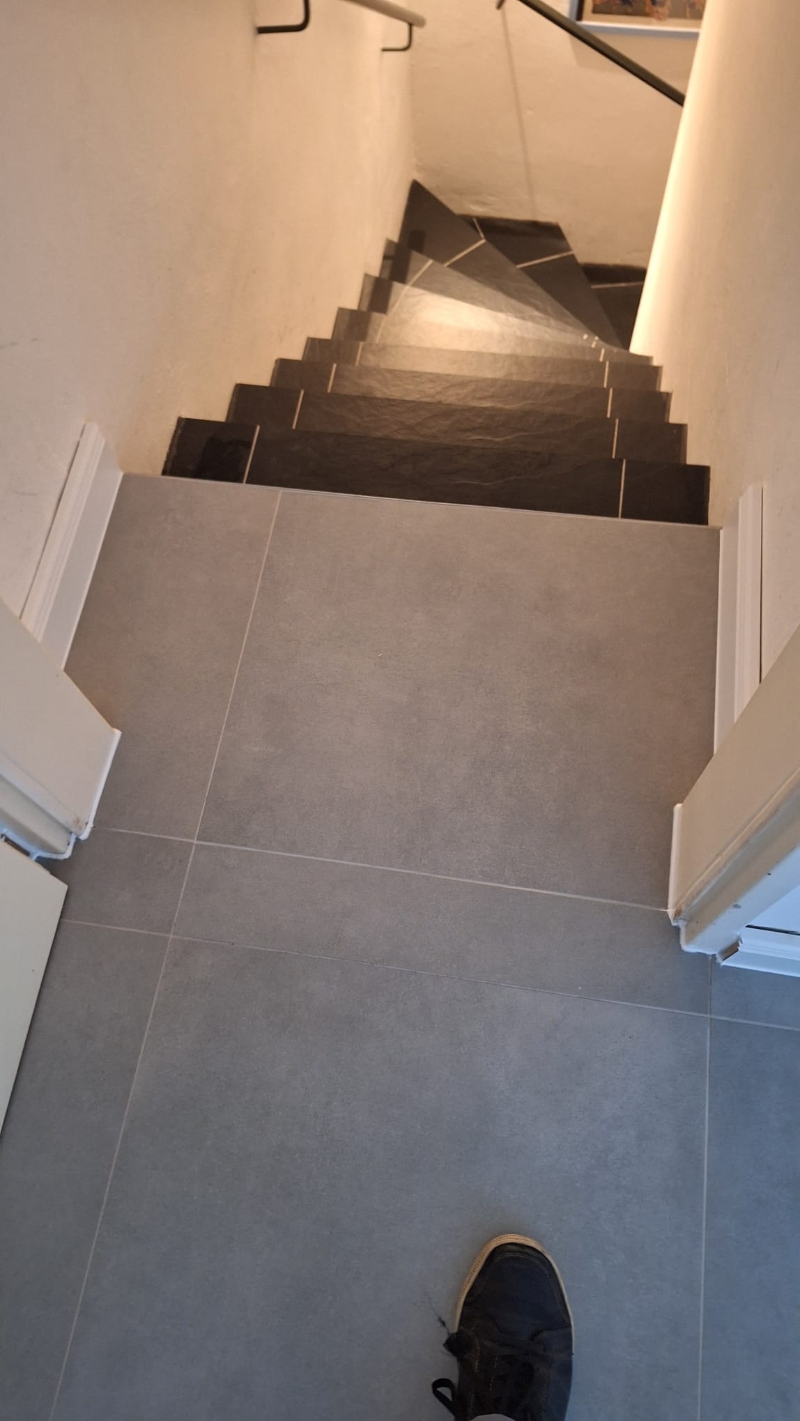 Szare płytki podłogowe i ciemne płytki schodowe, widok z góry. Widoczne fragmenty drzwi po bokach i but w rogu. Minimalistyczny design.