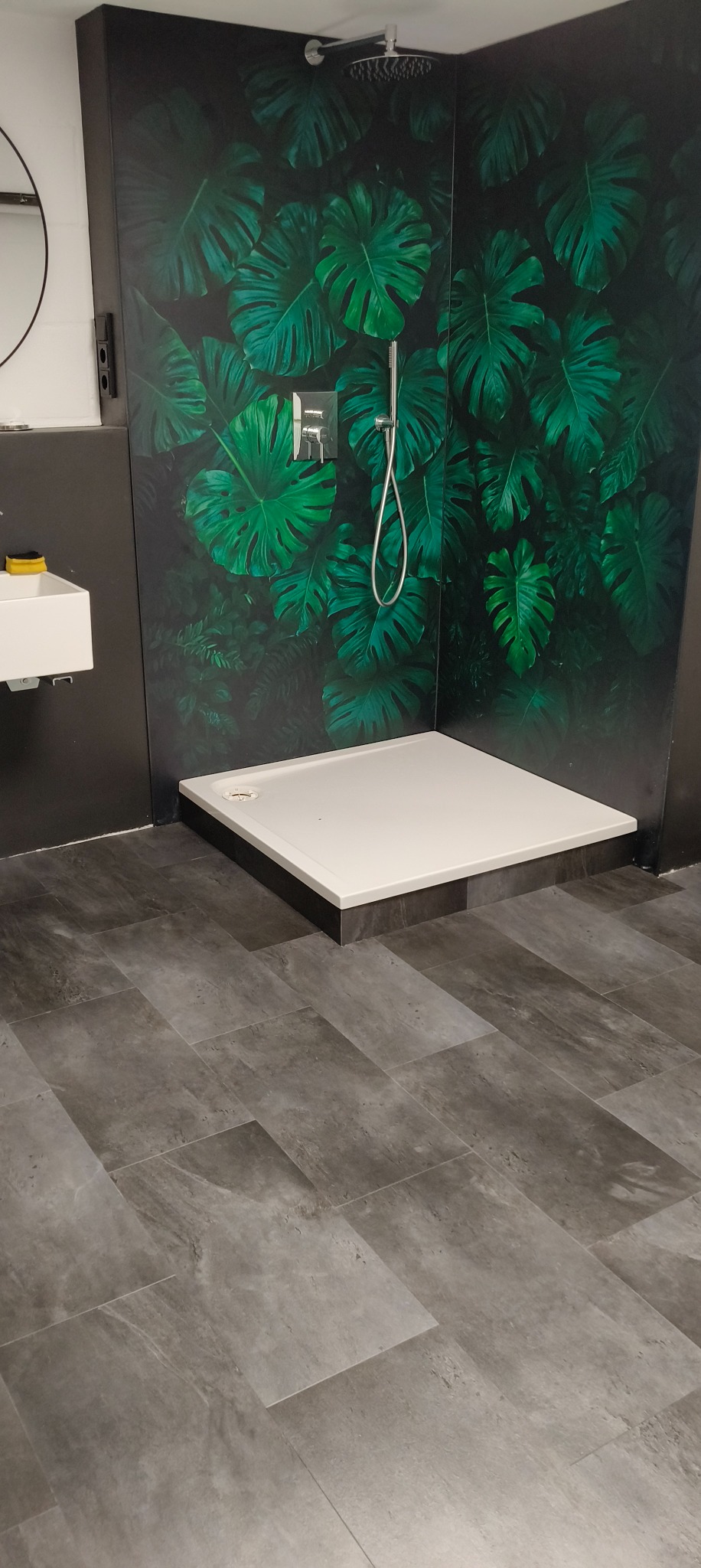 Nowoczesna kabina prysznicowa z tropikalnym motywem liści na ścianie, szara podłoga imitująca kamień, biały brodzik i chromowana armatura.