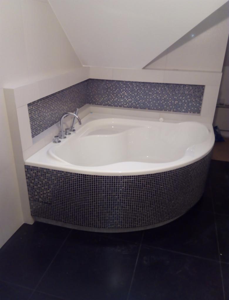 Narożna wanna obudowana mozaiką w odcieniach szarości i błękitu, z baterią umieszczoną na obudowie, na tle czarnej podłogi i białych ścian ze skosem.