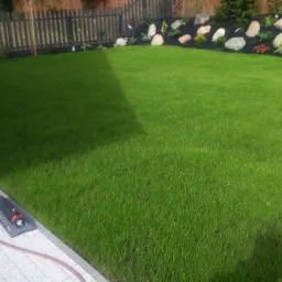 Świeżo skoszony trawnik z systemem nawadniania w postaci zraszacza oscylacyjnego leżącego na jasnej kostce brukowej, obok węża ogrodowego. W tle widoczny ciemny płot i rabata z kamieniami i roślinami.