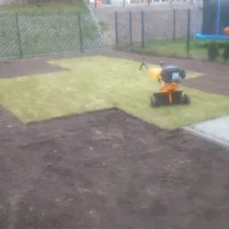 Układanie trawy z rolki w Gdańsku, widoczny traktor glebogryzarka na częściowo ułożonym trawniku, w tle ogrodzenie z siatki i trampolina.