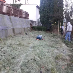 Dwaj mężczyźni w Gdańsku sadzą młode drzewko, opierając je o drewniane paliki. Na pierwszym planie zroszona trawa, w tle ogrodzenie i budynek.