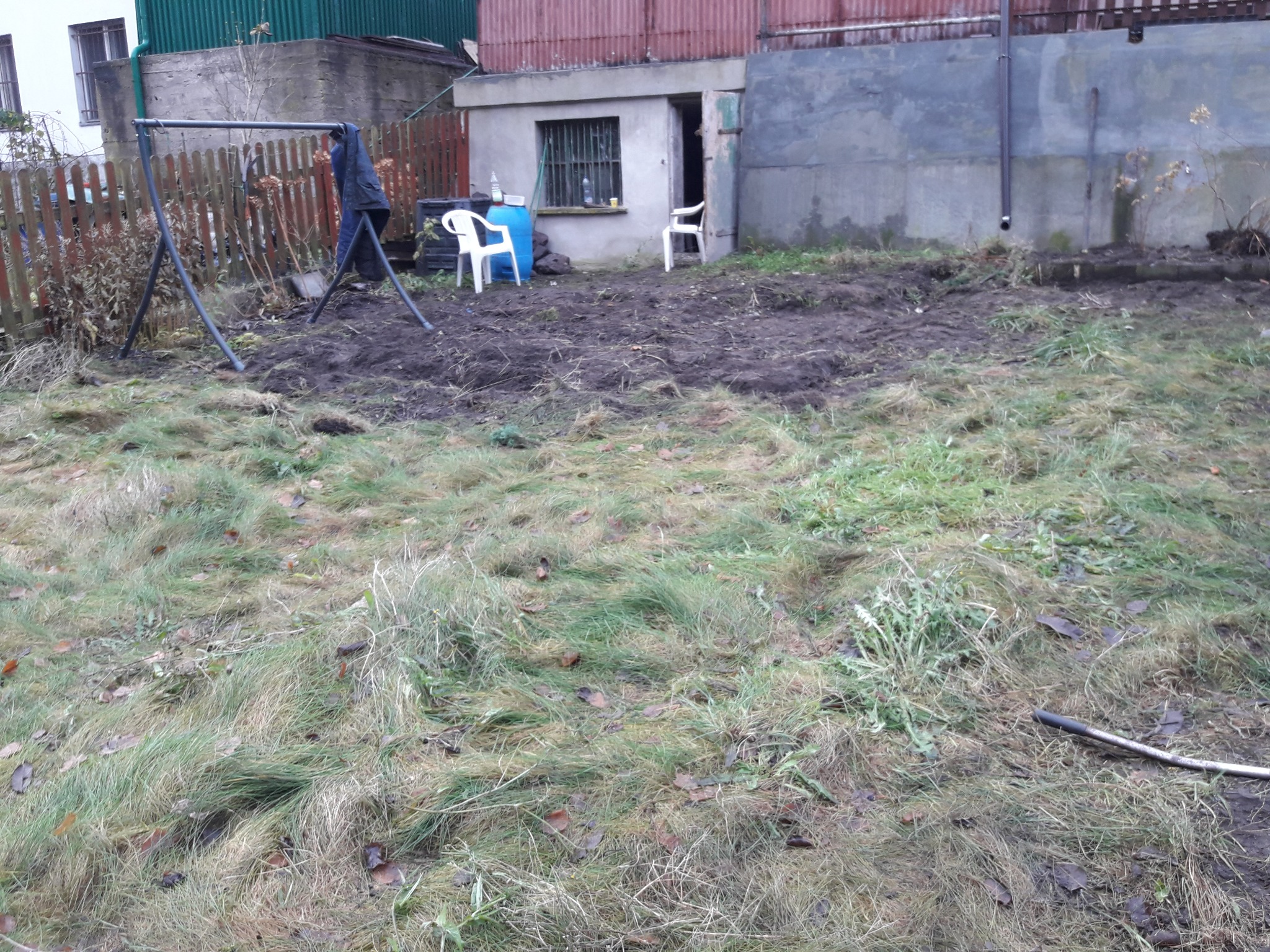 Przygotowany teren pod ogród z przekopaną ziemią, widoczne pozostałości trawy, w tle budynek gospodarczy i huśtawka ogrodowa.
