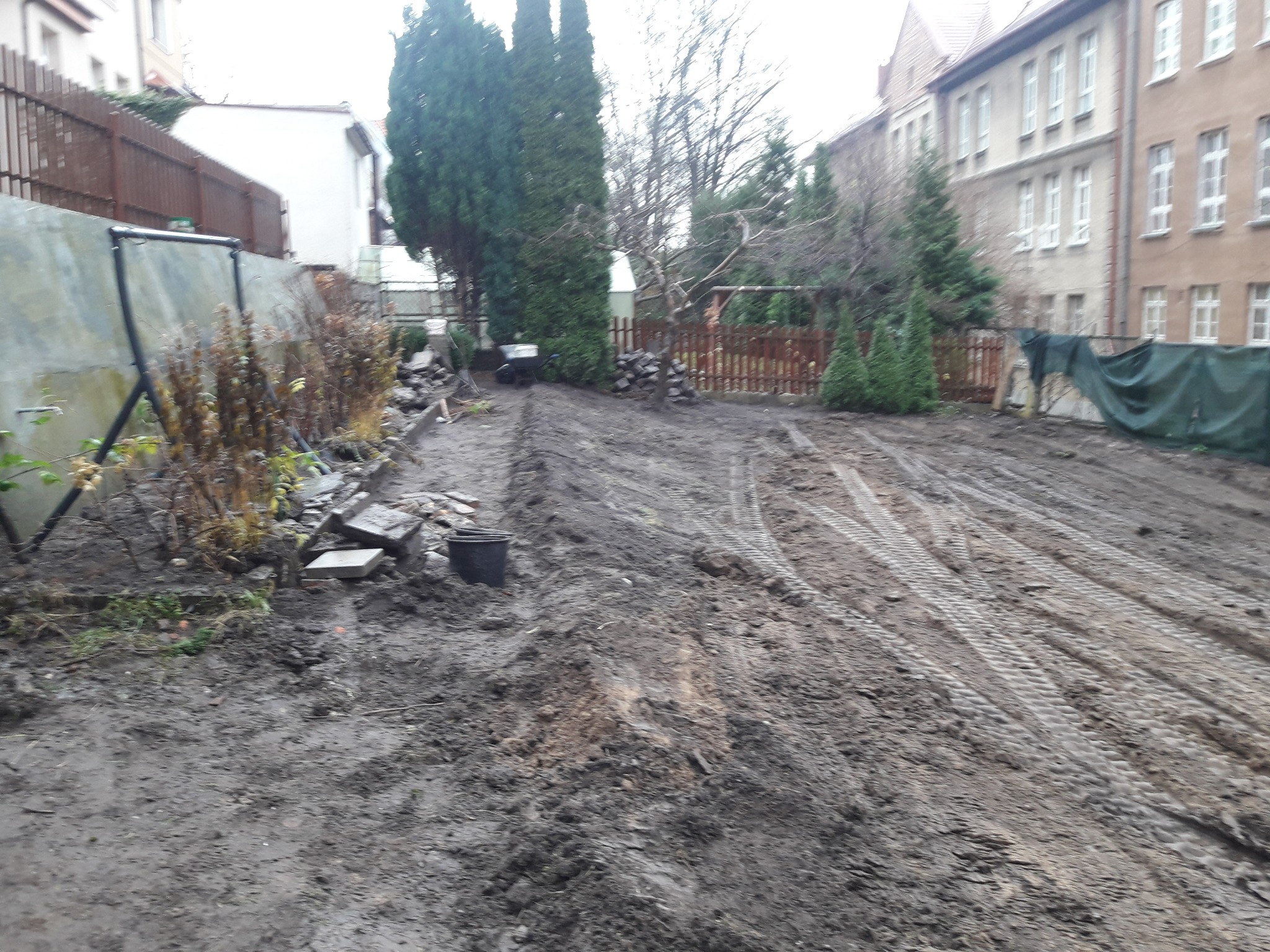 Przygotowany teren pod ogród z widocznymi śladami opon, częściowo usuniętą roślinnością i elementami dekoracyjnymi, takimi jak kamienie i doniczka, w tle budynki mieszkalne i drzewa.