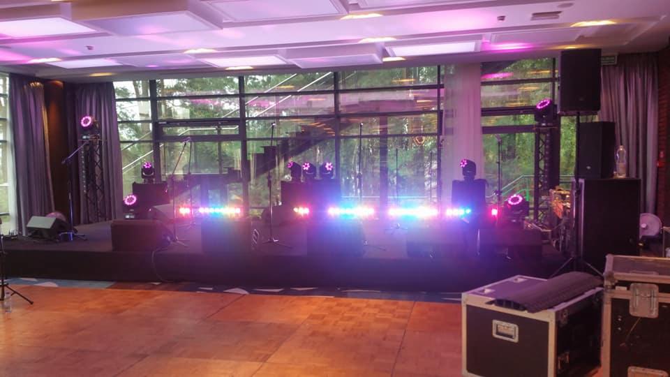 Scena z nagłośnieniem i oświetleniem przygotowana do występu, widoczne mikrofony, kolumny głośnikowe, reflektory i skrzynie transportowe, tło stanowią okna z widokiem na zieleń.