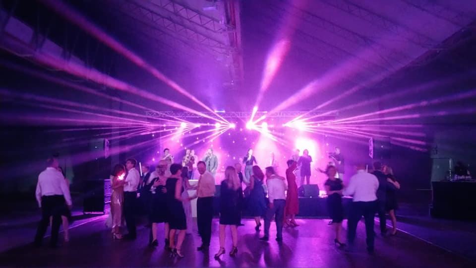 Zespół muzyczny na scenie podczas imprezy, oświetlony fioletowymi laserami i reflektorami, tańczący tłum w tle.