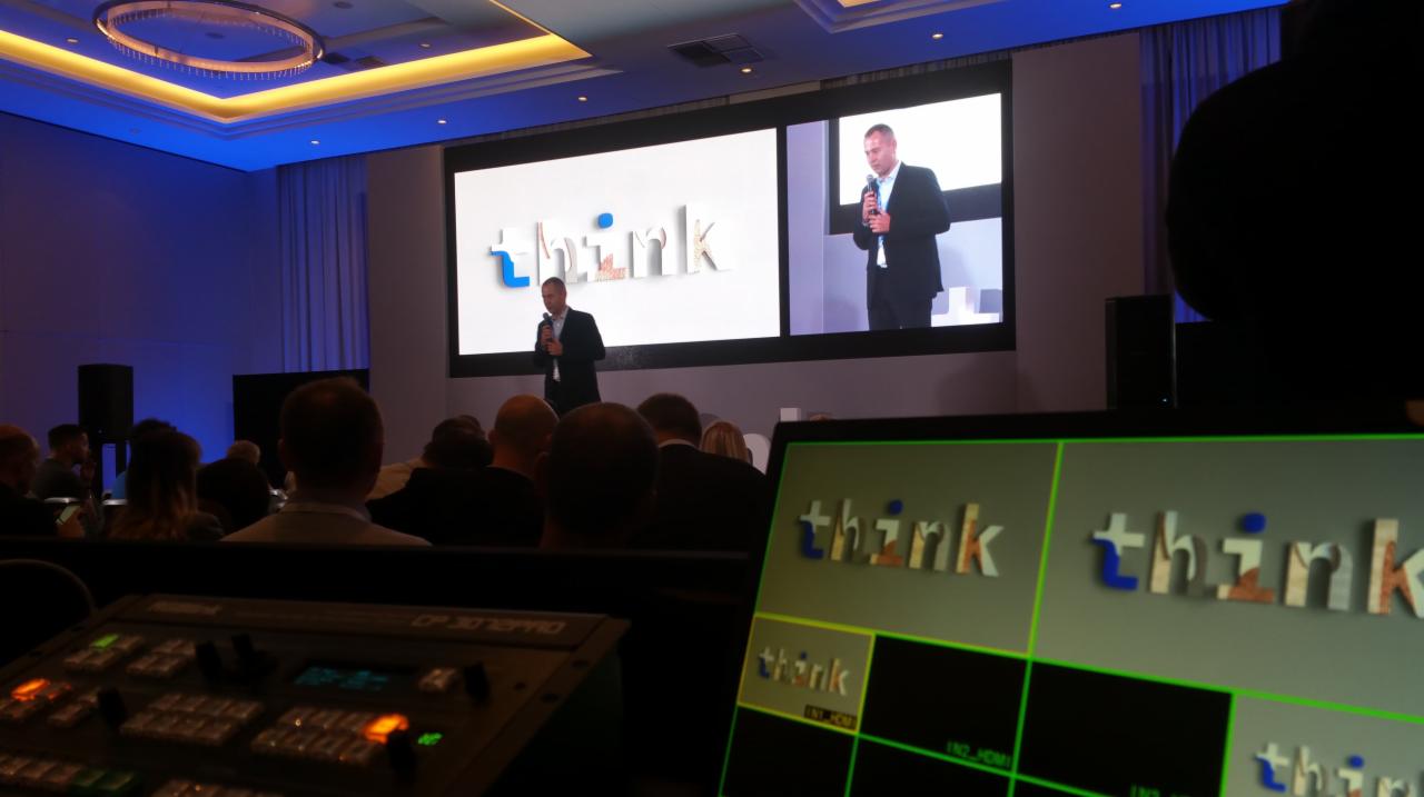 Prelegent w garniturze przemawia na scenie z dużym ekranem wyświetlającym logo 'think', widok zza konsolety realizatora z podglądem na żywo.