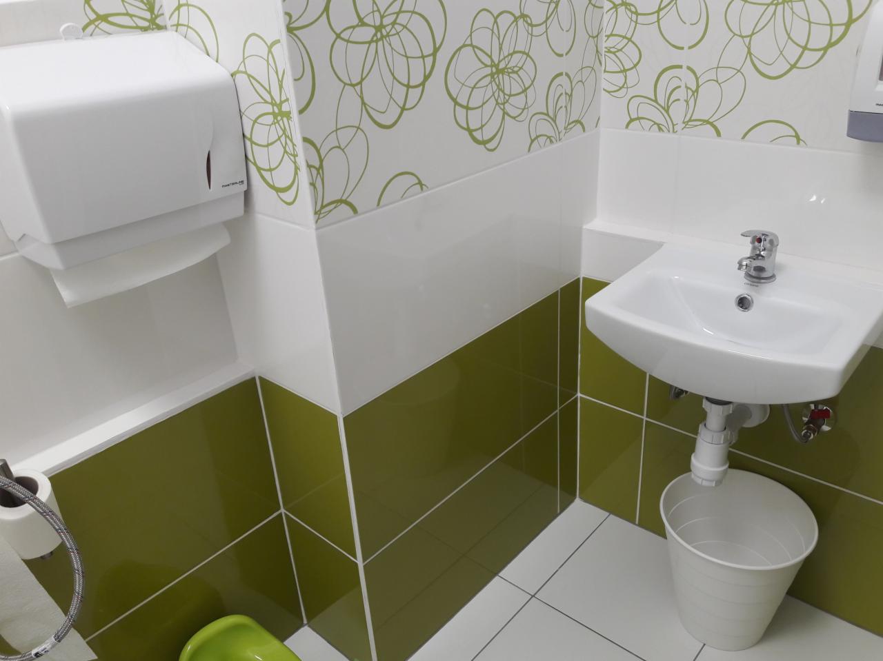 Umywalka z baterią i koszem pod spodem w toalecie wykończonej białymi i oliwkowymi płytkami, z dozownikiem ręczników papierowych i wzorem kwiatowym na ścianie.