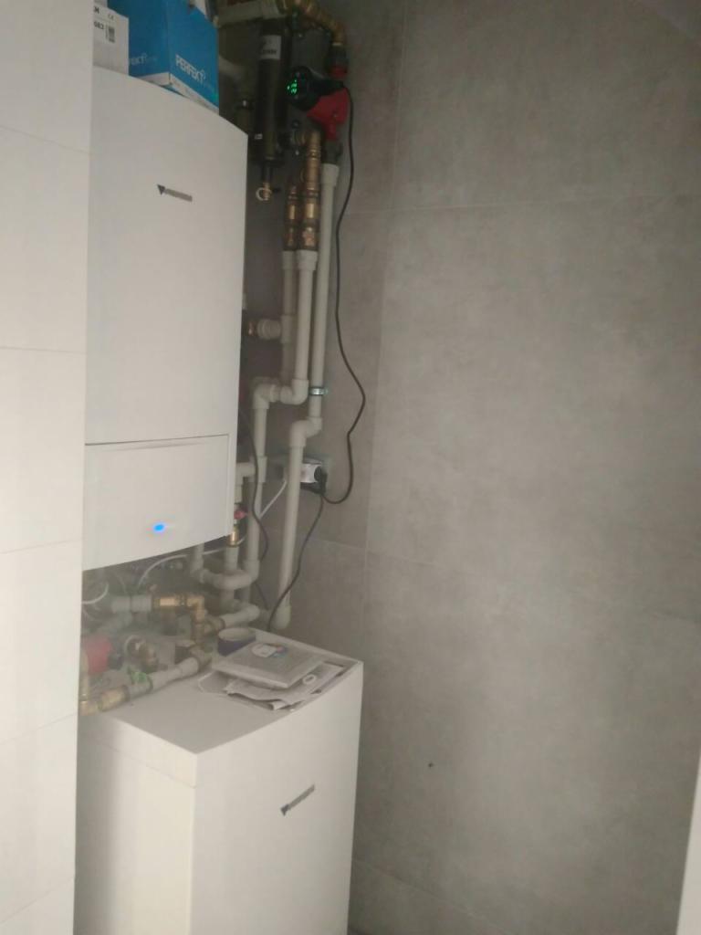 Biały piec grzewczy z licznymi rurami miedzianymi i plastikowymi, zainstalowany na tle szarych płytek ściennych. Na górze pieca leżą dokumenty.