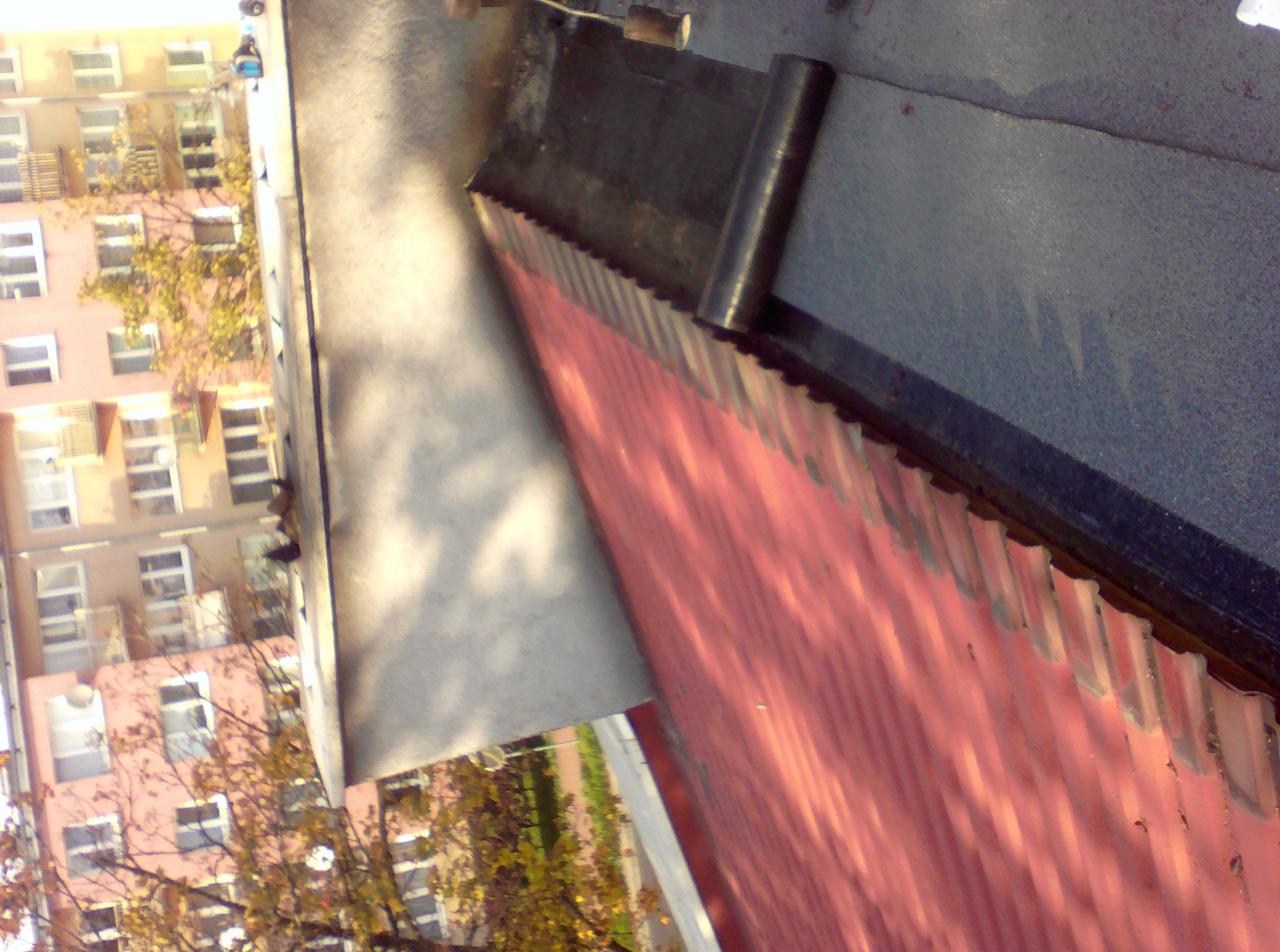 Fragment dachu z blachy trapezowej w kolorze czerwonym, z widocznym fragmentem papy bitumicznej i obróbki blacharskiej, na tle budynku mieszkalnego.