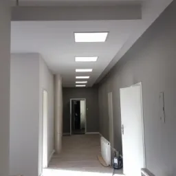 Korytarz biurowy po remoncie, ściany pomalowane na szaro, sufit z wbudowanymi panelami LED, widoczne drzwi i grzejnik.