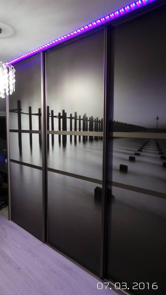 Szafa przesuwna z fototapetą przedstawiającą molo nad morzem, oświetlona fioletowymi diodami LED u góry.