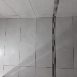 Wykończona ściana w łazience z jasnoszarych, matowych płytek ceramicznych z dekoracyjnym pionowym pasem mozaiki w odcieniach szarości.