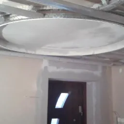 Okrągła konstrukcja sufitowa z płyty gipsowo-kartonowej, zamontowana na metalowym stelażu, w trakcie prac wykończeniowych, widoczne ślady szpachlowania na ścianach i nadprożach.