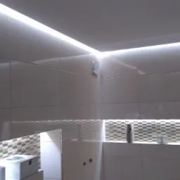 Jasne płytki w łazience z oświetleniem LED podsufitowym i dekoracyjnym pasem mozaiki, widoczna wentylacja i akcesoria łazienkowe.