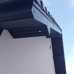 Fragment elewacji budynku z widocznym, ciemnym podbitkiem dachowym i oświetleniem punktowym w nim zamontowanym, na tle jasnego tynku elewacyjnego i błękitnego nieba.