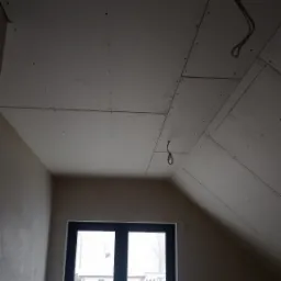 Skośny sufit poddasza wykończony płytami gipsowo-kartonowymi, widoczne łączenia i wkręty, z wiszącymi kablami elektrycznymi, w tle okno z widokiem na zaśnieżony ogród.