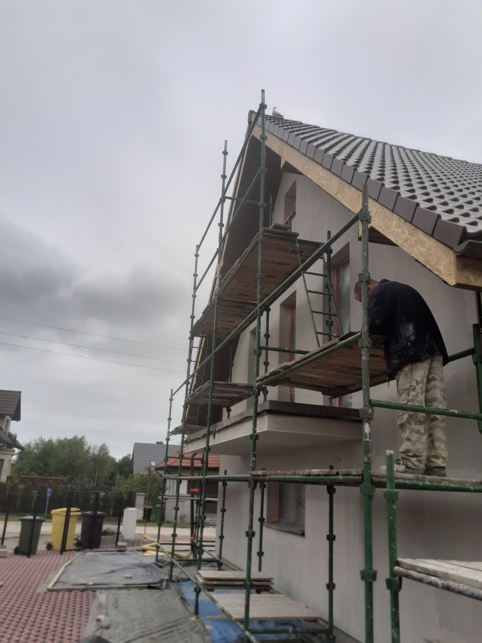 Pracownik na rusztowaniu, w trakcie nakładania warstwy wykończeniowej na elewacji domu jednorodzinnego z brązową dachówką, widoczne okna zabezpieczone folią, pochmurne niebo.