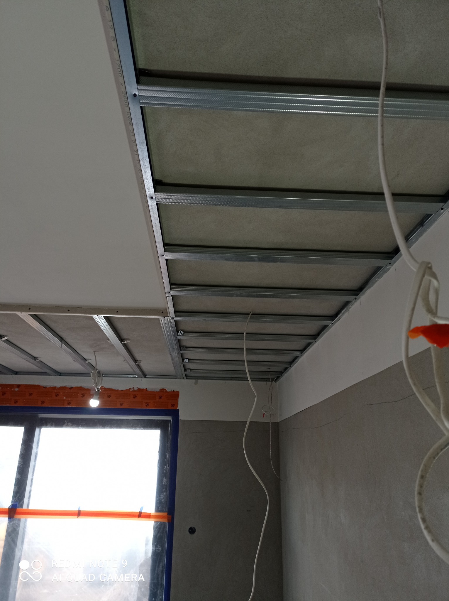 Montaż sufitu podwieszanego: widoczne metalowe profile konstrukcyjne i częściowo zamontowane płyty, okno zabezpieczone folią malarską w tle, zwisające kable elektryczne.