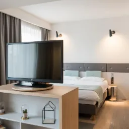 Nowoczesny pokój hotelowy z telewizorem LG na drewnianej szafce, łóżkiem z szarą ramą i zagłówkiem, oraz biurkiem z lampką, oświetlony dwoma kinkietami na białej ścianie.