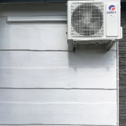 Biała jednostka zewnętrzna klimatyzacji marki Gree zamontowana na jasnej, drewnianej elewacji budynku z elementami dekoracyjnymi z ciemnego kamienia.