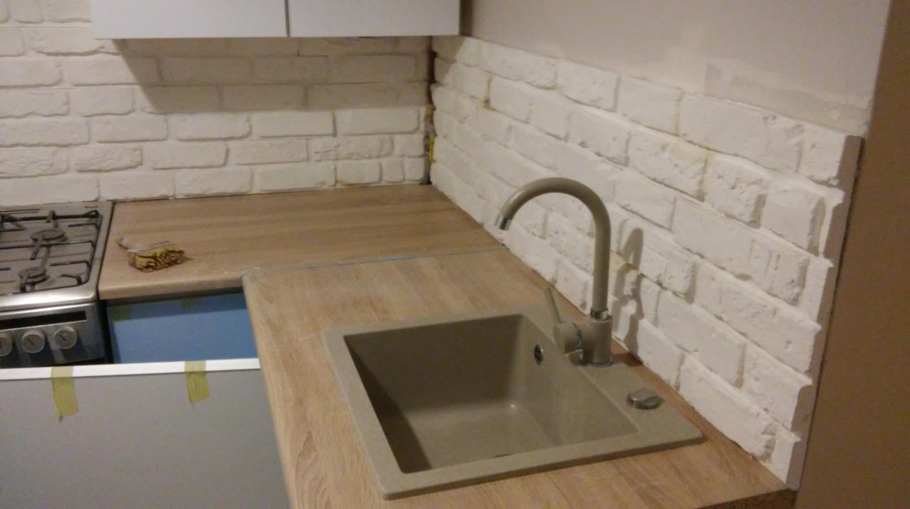 Kuchenny blat z drewnianym wzorem, z beżowym zlewem i baterią, ściana wyłożona białymi cegiełkami, fragment kuchenki gazowej po lewej stronie.