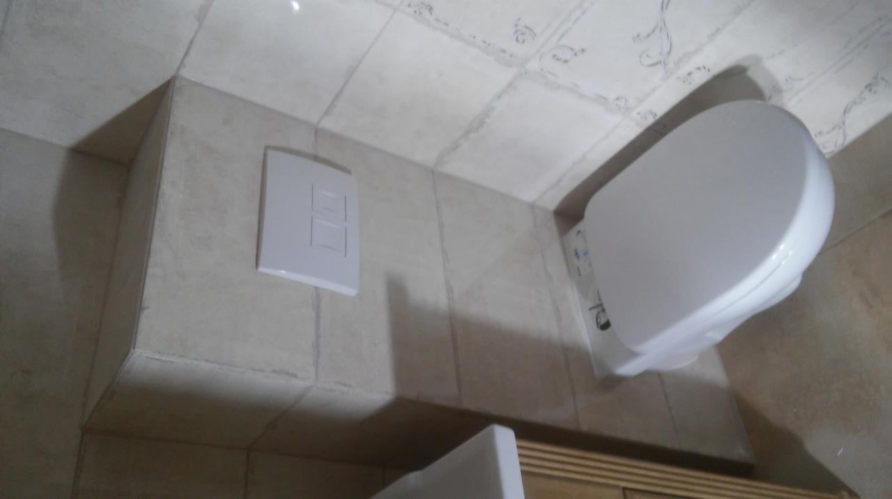 Świeżo wykończona toaleta podwieszana z beżowymi płytkami i podtynkowym systemem spłukiwania z białym przyciskiem. Widoczne niedoskonałości w fugowaniu płytek.