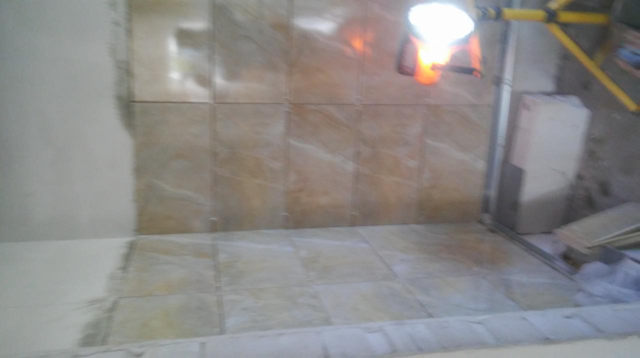 Ściana i podłoga w trakcie kładzenia płytek imitujących marmur, widoczna lampa budowlana i fragment ściany przygotowanej do dalszych prac wykończeniowych.