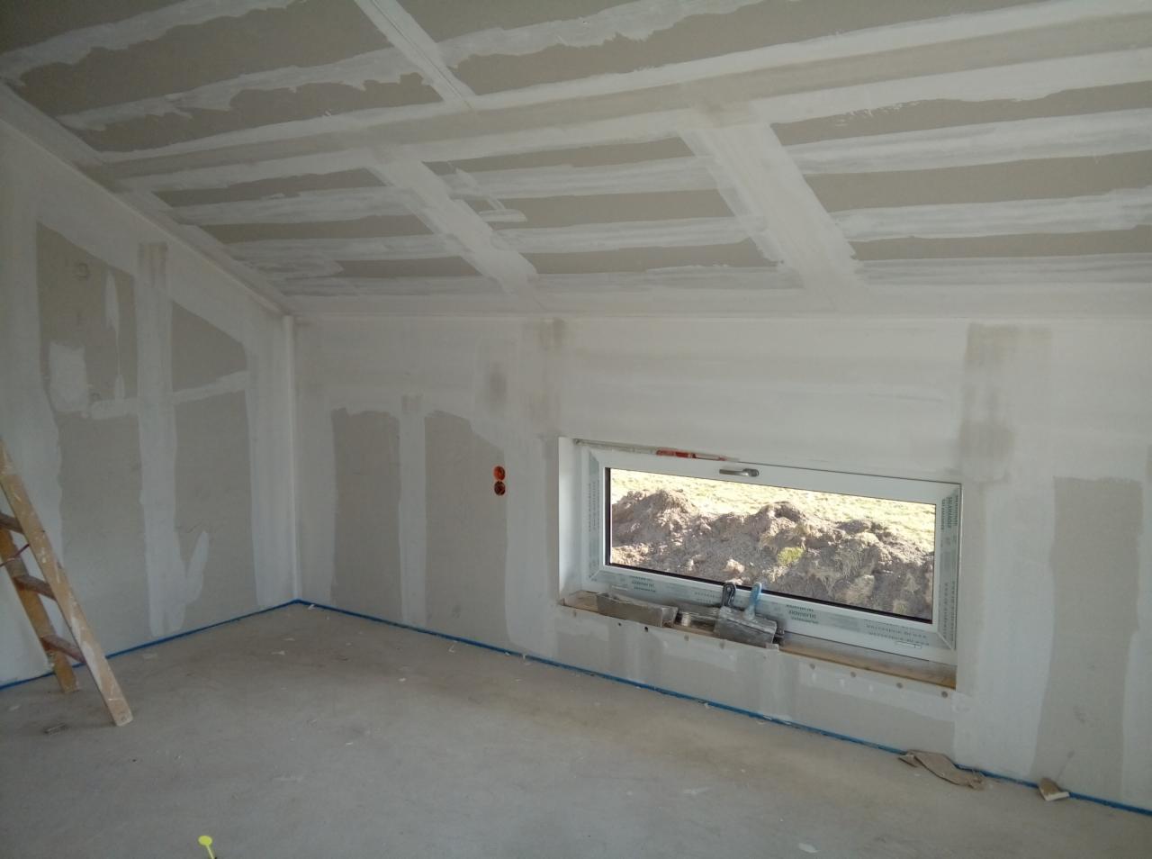 Poddasze w trakcie prac wykończeniowych: ściany i sufit pokryte płytami gipsowo-kartonowymi, widoczne łączenia płyt wzmocnione taśmą, okno z widokiem na ziemię, drabina oparta o ścianę, niebieska...