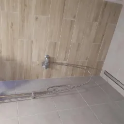 Wykończona łazienka z drewnopodobnymi płytkami ściennymi, szarą podłogą i zamontowanym odpływem liniowym oraz baterią prysznicową z wężem.