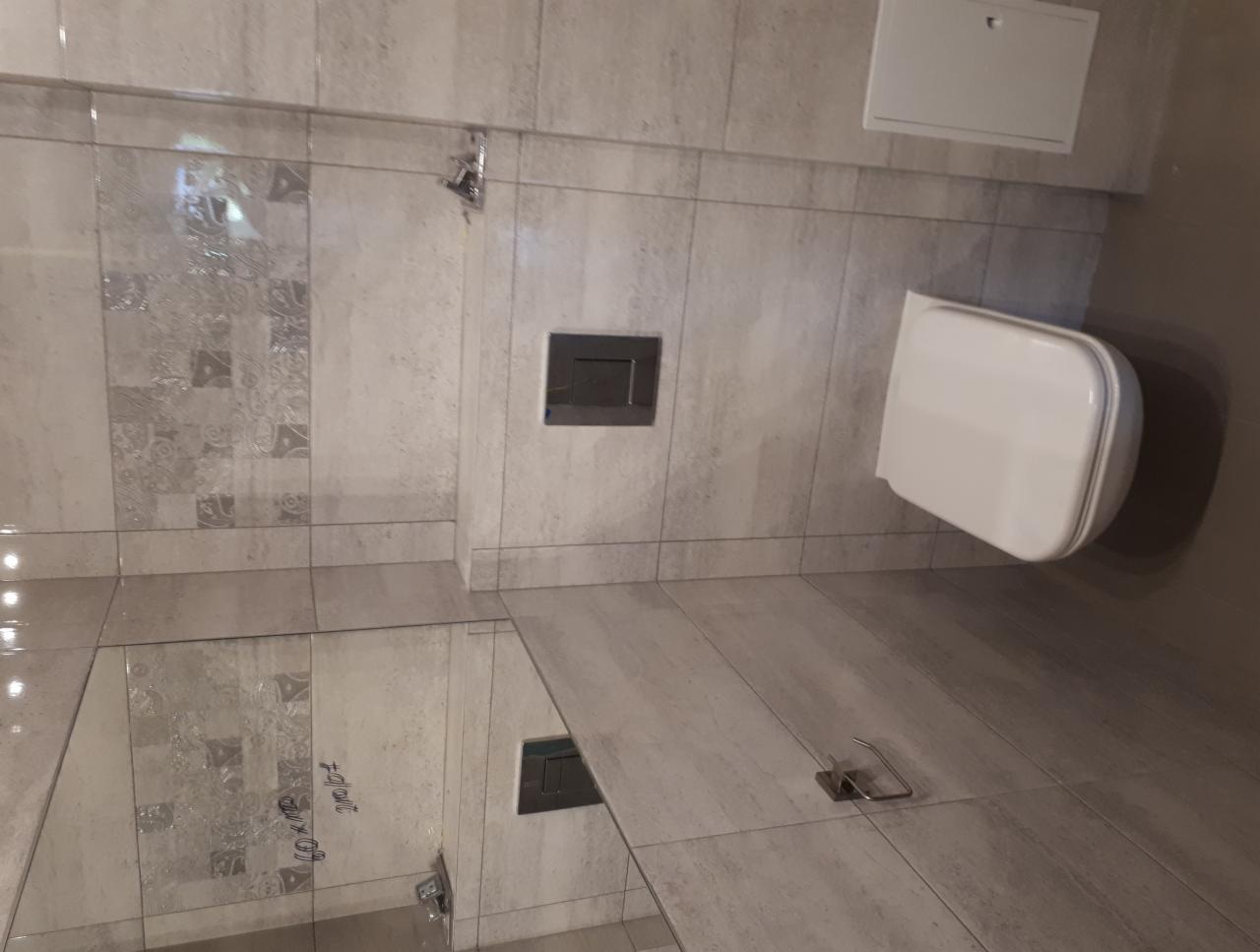 Nowoczesna toaleta wisząca na ścianie z szarymi płytkami z delikatnym wzorem, widoczny przycisk spłukiwania i uchwyt na papier toaletowy, odświeżona łazienka