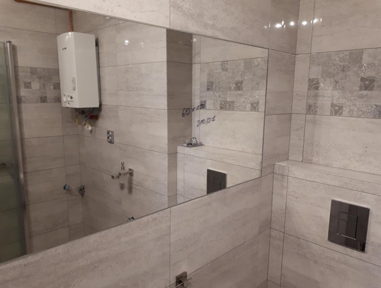 Wnętrze łazienki z beżowymi płytkami imitującymi kamień, widoczne lustro z odblaskiem, bojler oraz przyciski spłuczki. Na lustrze widoczne notatki pisane markerem.