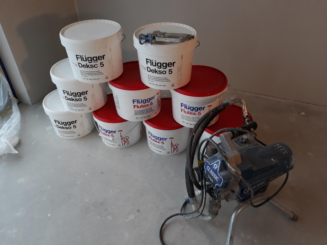 Stos wiader z farbami Flugger Dekso 5 i Flutex-5 oraz agregat malarski Graco Classic 395 na szarym betonowym podłożu w rogu pomieszczenia.