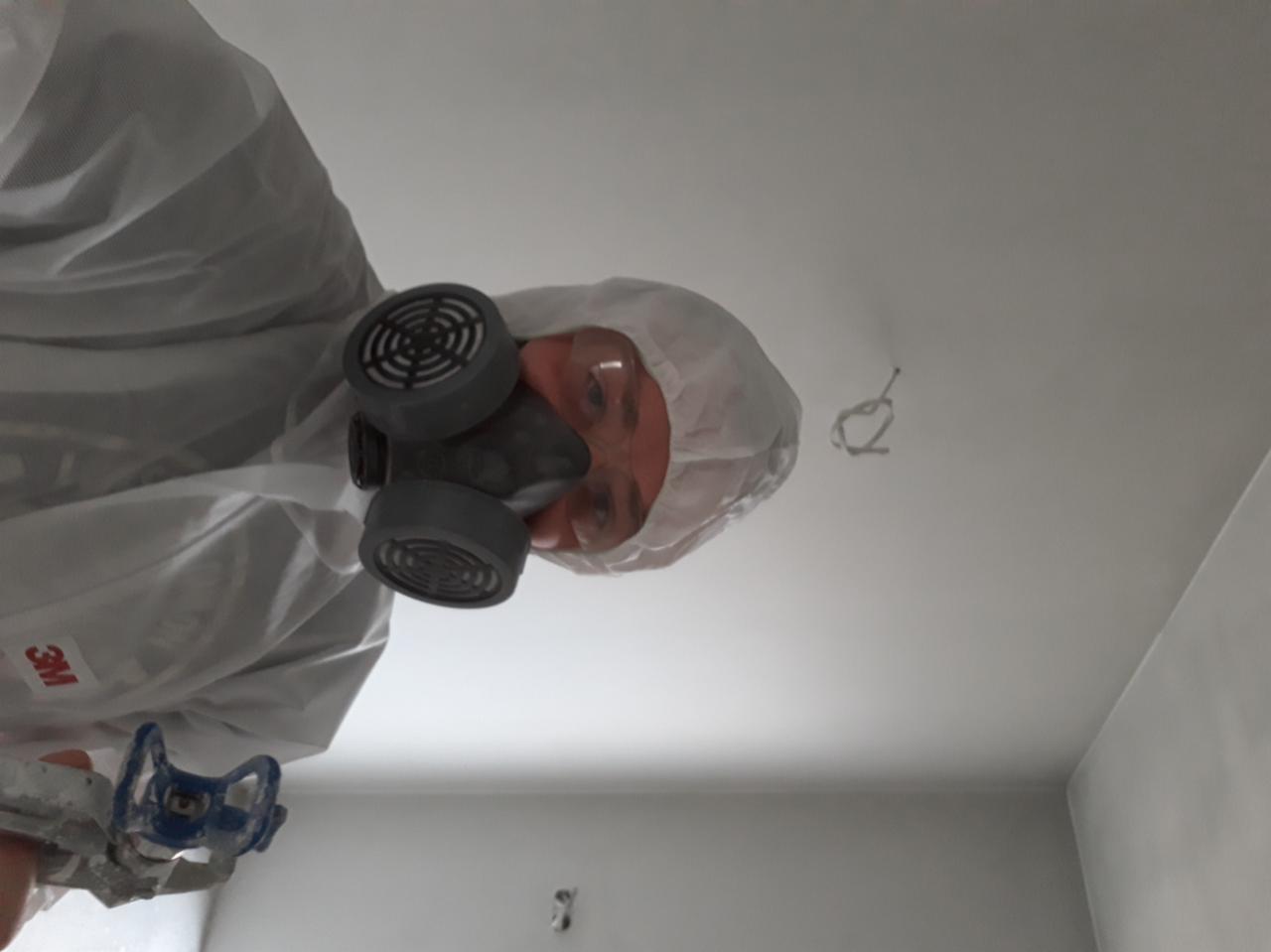 Pracownik w kombinezonie ochronnym i masce malarskiej, trzymający pistolet natryskowy, maluje sufit na biało. Widoczne wystające kable z sufitu.