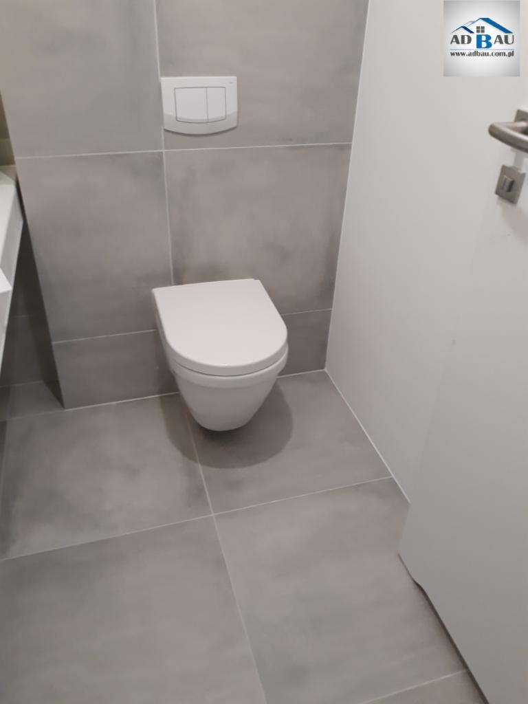 Szara łazienka z podwieszaną toaletą i dużymi płytkami podłogowymi, widoczne logo firmy AD BAU w prawym górnym rogu.
