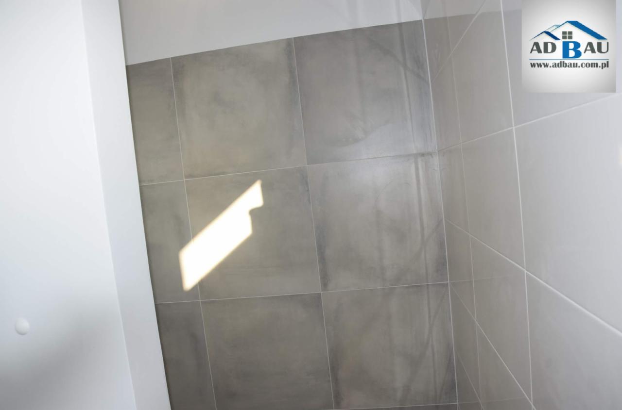Szara, kwadratowa glazura ścienna w łazience, oświetlona promieniem słońca, z widocznym logo firmy AD BAU w prawym górnym rogu.