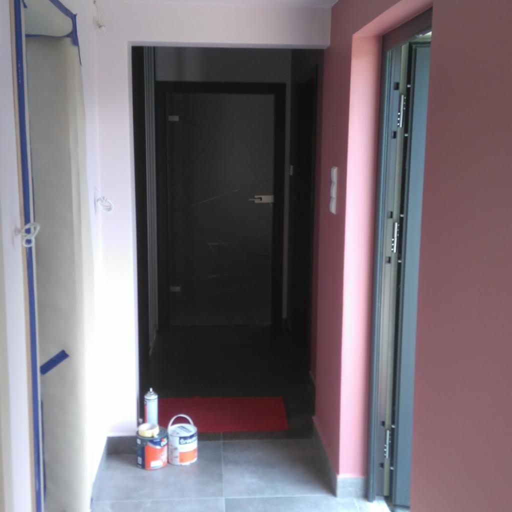 Korytarz w trakcie odświeżania z widocznymi puszkami farb i wałkiem, ściany pomalowane na różowo i biało, drzwi w kolorze ciemnego grafitu.