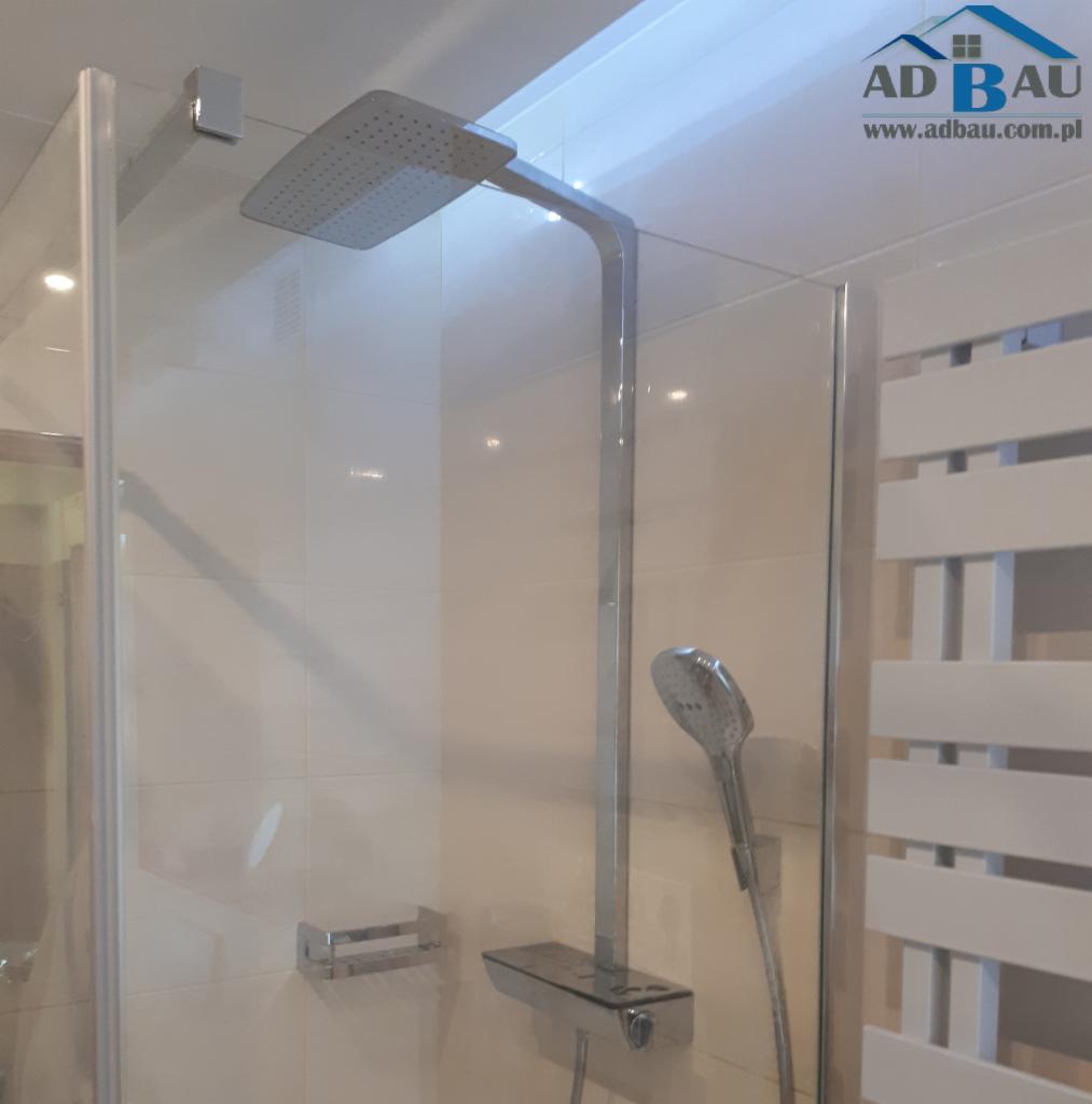 Nowoczesna kabina prysznicowa z deszczownicą i baterią termostatyczną, widoczny grzejnik drabinkowy oraz logo firmy AD BAU w rogu.