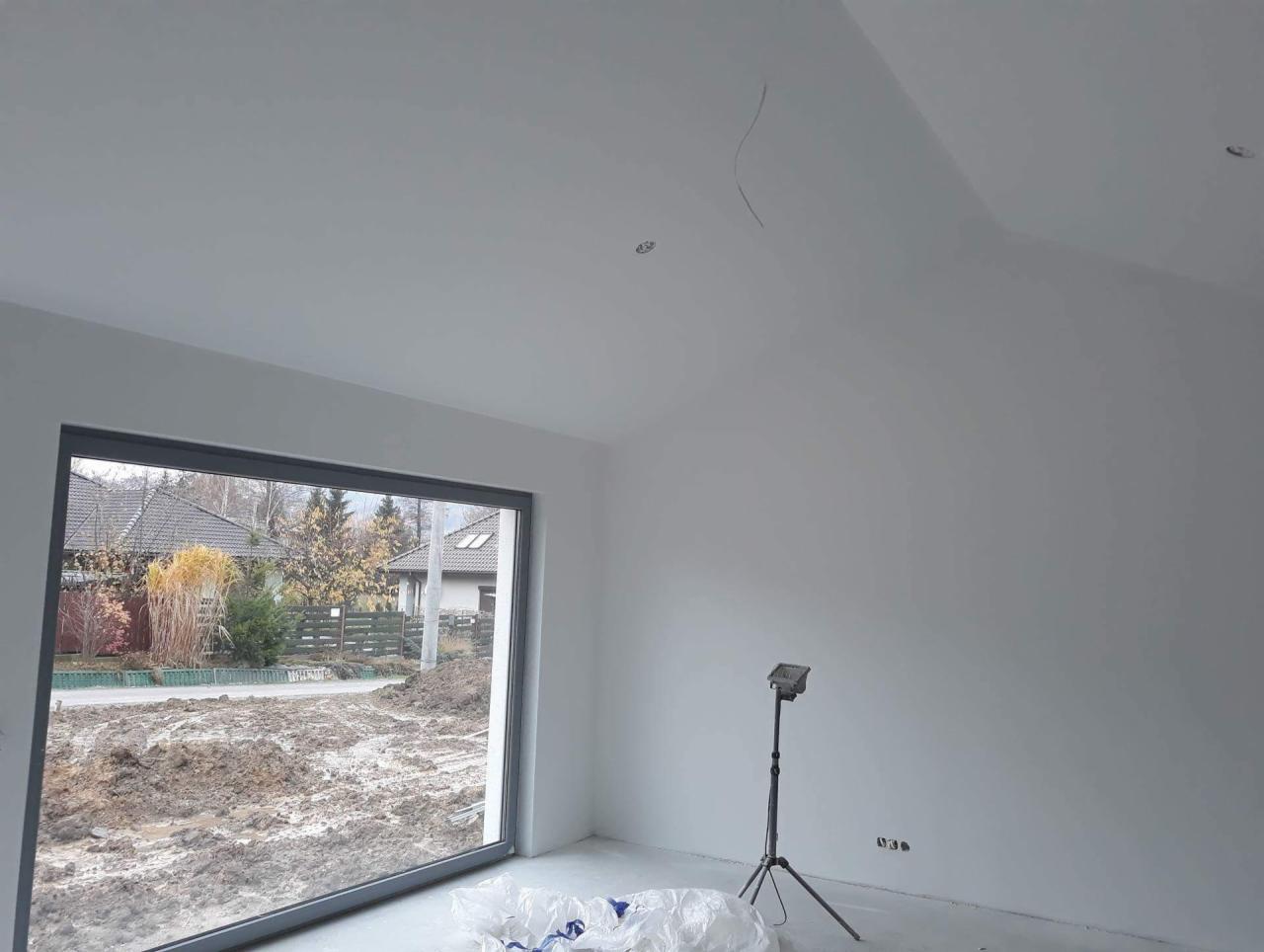 Wnętrze w trakcie odświeżania: świeżo pomalowane białe ściany, duże okno z widokiem na ogród z ziemią, lampa na trójnogu i folia ochronna na podłodze.
